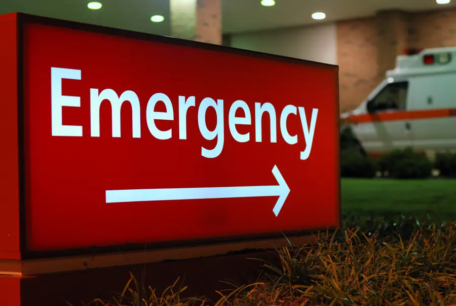 Illuminated red hospital emergency sign with an ambulance behind it